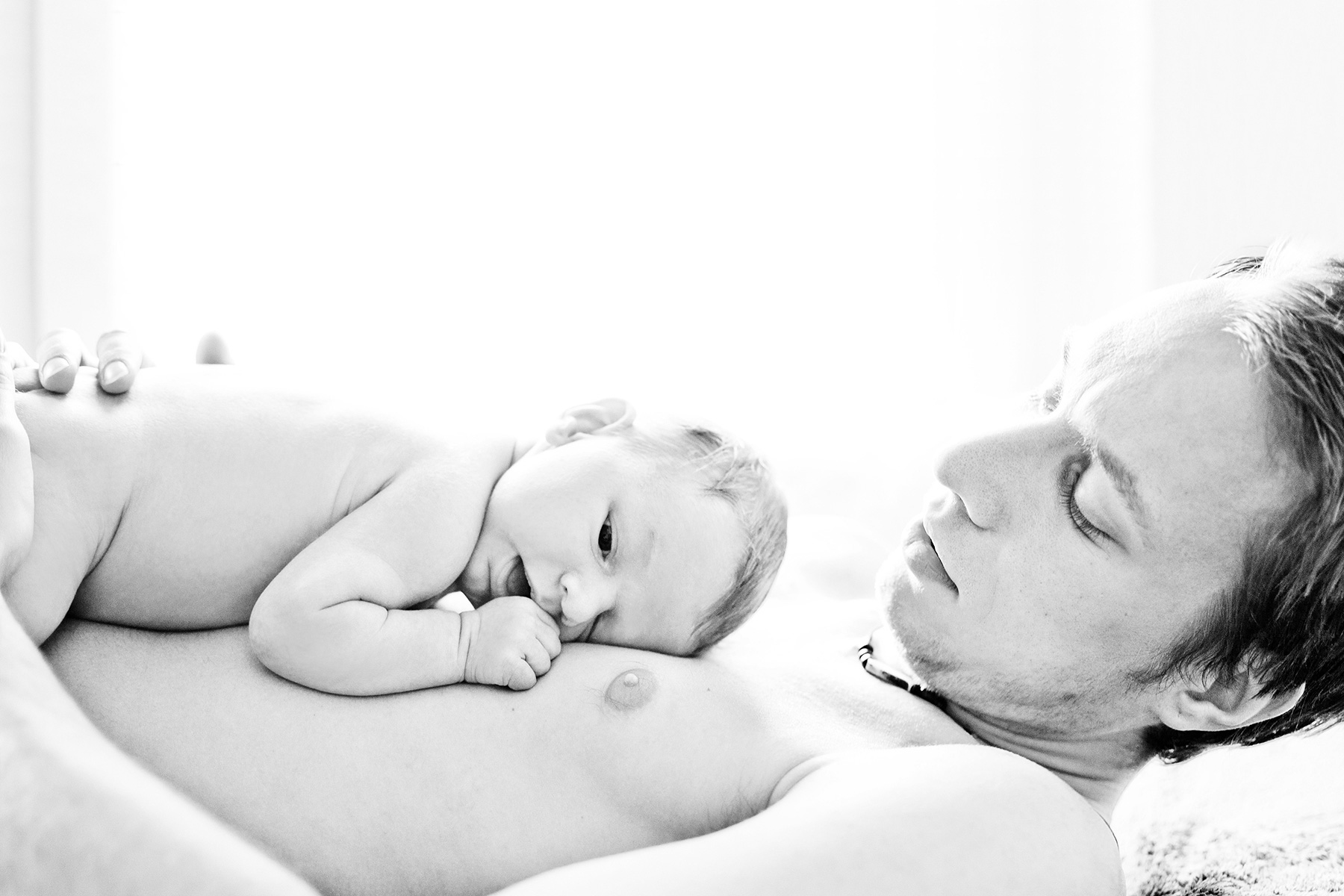 Papa liegt kuschelnd mit seinem Baby Bauch an Bauch auf dem Bett und blickt es verliebt an