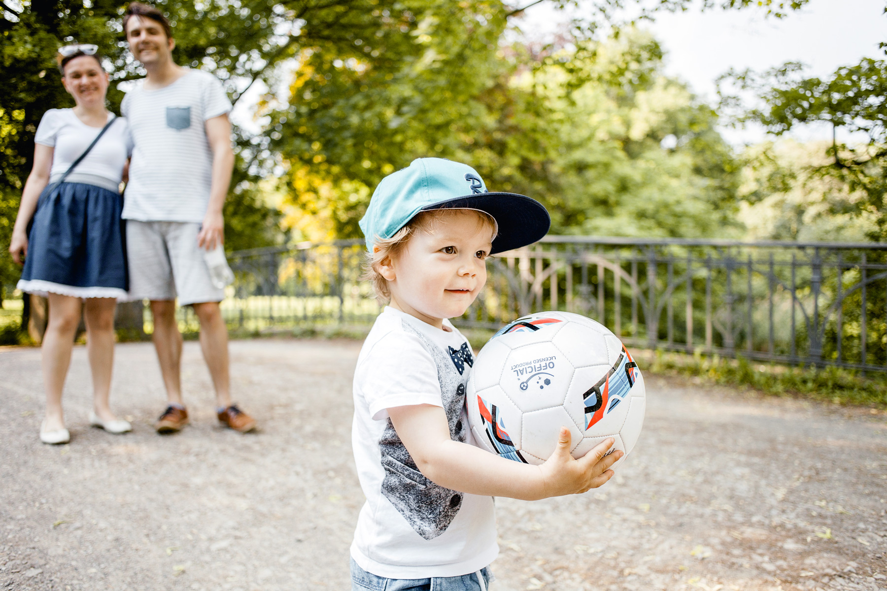 kleiner Junge steht mit seinem Fußball in den Händen stolz im Park. Eltern lächeln und beobachten ihn dabei.