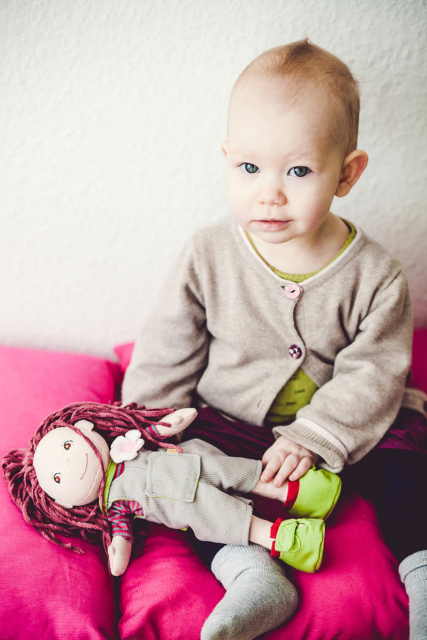 kleines Kind sitzt mit seiner Puppe auf pinkfarbenen Kissen